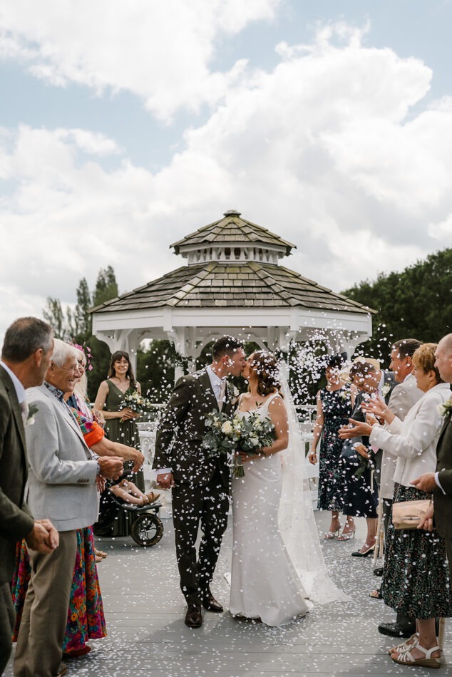 New husband and wife walking back down the aisle in a beautiful outdoor venue. Guests are throwing confetti as they walk past.